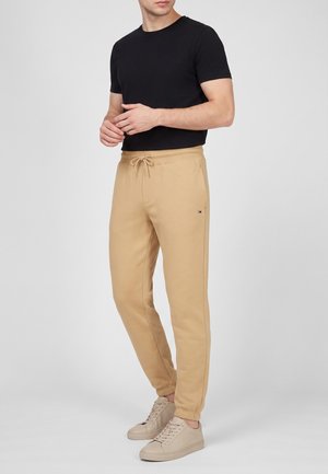 Uomo che indossa una maglietta nera a maniche corte, pantaloni da jogging beige con coulisse e sneakers beige, in piedi davanti a uno sfondo semplice.