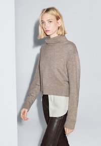 Sweter z golfem w jasnobrązowej wełnianej mieszance, skrócona konstrukcja, długie rękawy, noszony na białej koszuli z guzikami i ciemnych skórzanych spodniach.