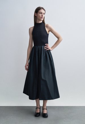 Mladá žena v čiernom bezrukávovom topu a širokej čiernej sukni po členky stojaca s jednou rukou na boku pred jednoduchým pozadím.