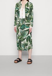 Vrouw in een groene rok met tropisch bladenprint en bijpassende ritsjack, witte top met uitsnede en zwarte sandalen met twee banden, staande voor een effen achtergrond.