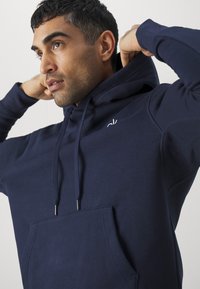 Marineblaues Kapuzensweatshirt mit Kängurutasche, Kordelzügen mit metallischen Enden und einem kleinen Logo auf der Brust. Weicher Stoff.