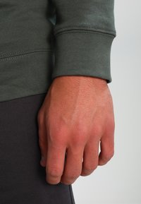 Sweatshirt verde a maniche lunghe con polsini a costine; una mano posizionata al polso, che mostra il tessuto testurizzato e pantaloni grigio scuro sottostanti.