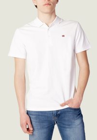 Polo blanc en coton avec une texture subtile, à manches courtes, et un petit logo rouge et bleu sur le côté gauche de la poitrine. Associé à un jean bleu.
