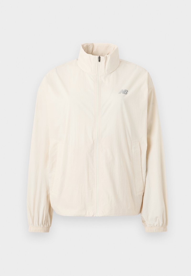 Veste légère de couleur crème avec un col montant, fermeture éclair frontale complète, poignets élastiques et un petit logo gris en haut à gauche.