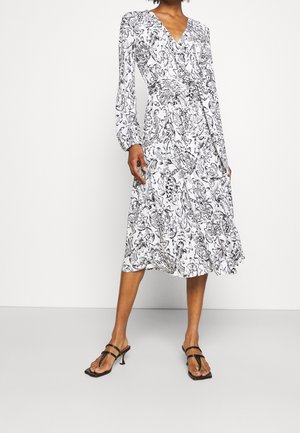 Kvinde iført en hvid midi-kjole med indviklede sorte blomsterprint og lange ærmer, kombineret med sorte sandaler med hæl og stropper.