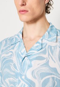 Camicia a maniche corte color celeste e bianca con un motivo a spiral, dotata di colletto a punta e tre bottoni sul davanti.