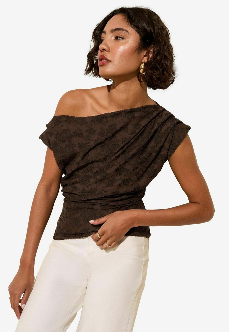 Bruine off-shoulder top met bloemstructuur, gedrapeerd ontwerp en korte mouwen, gecombineerd met lichtgekleurde broek.