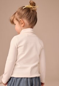 Long-sleeve cream top with a high collar, paired with a blue pleated skirt. Hair styled in a bun with a light yellow ribbon accent.