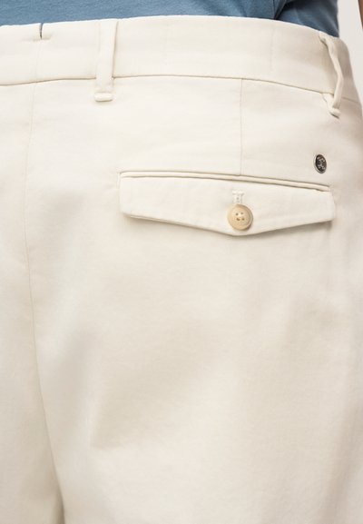 Μπεζ βαμβακερό παντελόνι με μπροστινή τσέπη που διαθέτει ένα ανοικτό καφέ κουμπί. Το ύφασμα έχει λεία υφή και είναι ελαφρώς τσαλακωμένο.