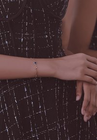 Bracciale in oro con dettaglio di pietra preziosa blu, caratterizzato da un design a catena. Sullo sfondo si vede un outfit nero texture con cuciture bianche.