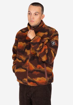 Mann trägt eine braun-orange-gelbe gemusterte Fleecejacke, mit einer Hand am Kinn und der anderen in der Tasche, schaut zur Seite.