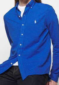 Blauwe langearmpolo met knopen en een polo-logo, met een opstaande kraag, witte knopen en een gestructureerde stofafwerking.