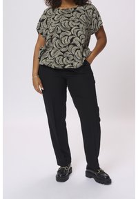 Frau mit schwarzen Hosen, schwarzen Loafers und einer schwarzen gemusterten Bluse mit kurzen Ärmeln, die mit einer Hand in der Tasche steht.