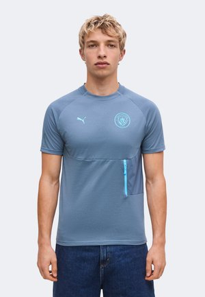 Jonge man draagt blauw Manchester City sportshirt met korte mouwen, Puma-logo en een ritszakje, staand tegen een effen lichte achtergrond.