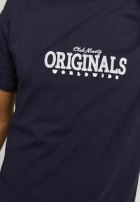 Camiseta de algodón azul marino con texto blanco: "Club Ninety ORIGINALS WORLDWIDE". Diseño de mangas cortas y cuello redondo.