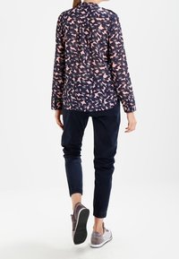 Kvinna i en rosa och svart blommig långärmad blus, marinblå uppvikta byxor och grå sneakers, som går bortåt på en vit bakgrund.