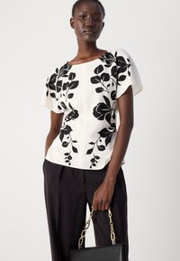 Witte top met zwart bloemenprint, voorzien van korte mouwen, textuurstof en een getailleerd ontwerp, gecombineerd met zwarte broek en een zwarte tas.