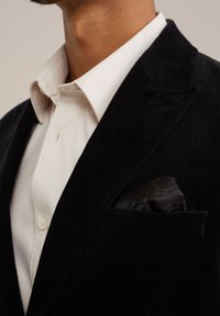 Zwarte corduroy blazer met notch-revers, witte dress shirt en een zwart gekleed zakdoekje zichtbaar in de borstzak.