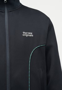 Fekete cipzáras dzseki fehér "the new originals" felirattal a mellrészen, jobb oldalon zseb körvonalán zöld kockás szegéllyel.