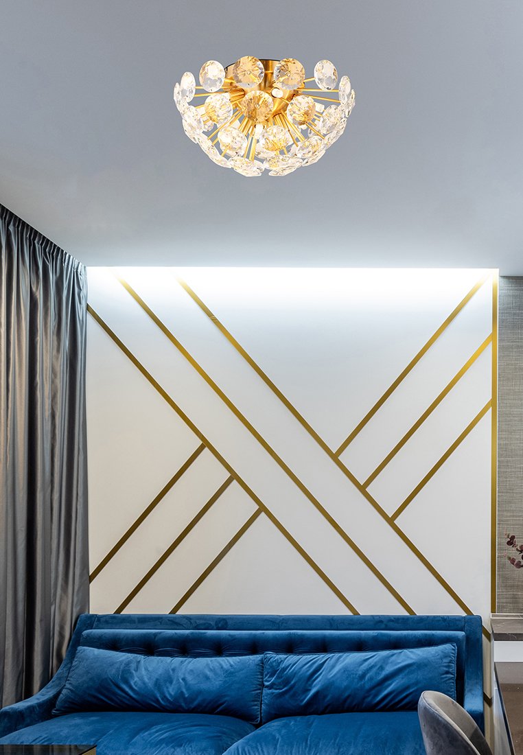 Kronlampa i kristall med en gyllene bas, prydd med cirkulära kristaller. Blå sammetssoffa mot en ljus vägg med diagonala gyllene linjer.