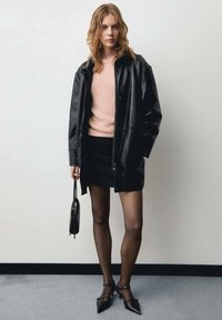 Crna kožna jakna preko ružičastog pulovera, uparena s crnom mini suknjom. Drži malu crnu torbicu, nosi prozirne čarape i cipele na špic.