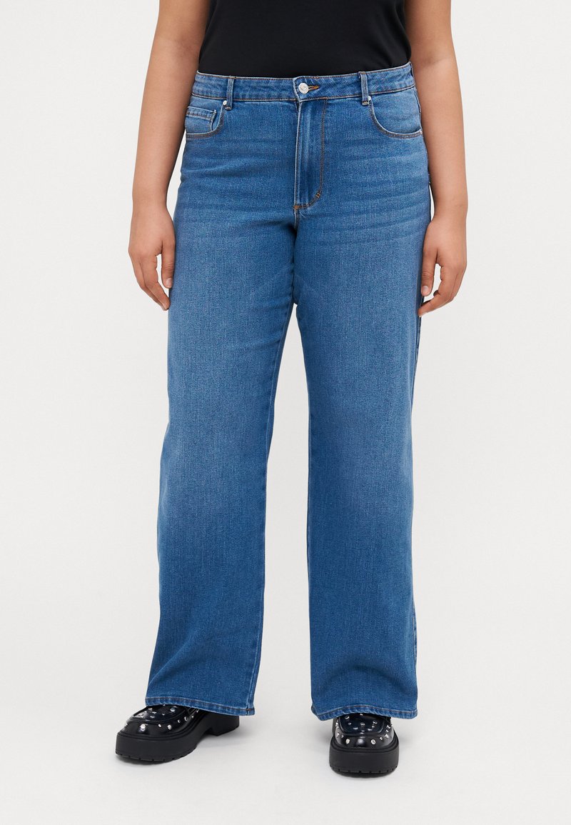 Wide-leg blue denim jeans with a high waist, five pockets, belt loops, and contrasting orange stitching. Paired with black platform shoes.
