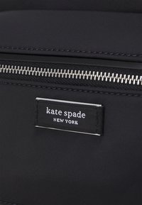Svart nylonmaterial med en slät yta har en silverfärgad dragkedja och en rektangulär Kate Spade-logotag i vit text.