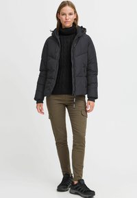 Doudoune noire avec une fermeture éclair, une capuche et un design matelassé, associée à un col roulé en tricot noir et à un pantalon cargo vert olive. Baskets noires.
