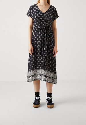 Kobieta w czarnej sukience midi z białymi wzorami w kształcie diamentów i szeroką wzorzystą bordiurą, w połączeniu z czarnymi skarpetkami i czarnymi trampkami.