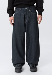 Person trägt dunkle Jeans mit weitem Bein, schwarze Turnschuhe und ein schwarzes Langarmshirt vor einem schlichten hellen Hintergrund.