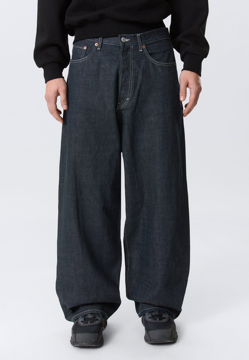 Person trägt dunkle Jeans mit weitem Bein, schwarze Turnschuhe und ein schwarzes Langarmshirt vor einem schlichten hellen Hintergrund.