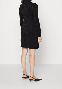 Schwarzes, geripptes Kleid mit langen Ärmeln und hohem Kragen, das eine figurbetonte Silhouette hat. Kombiniert mit schwarzen Slingback-Absätzen.