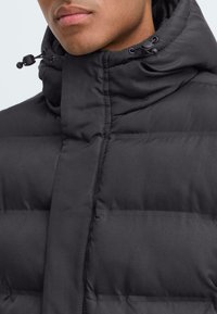 Chaqueta acolchada negra con un alto cuello, que presenta una textura acolchada y toggles ajustables en el cuello. Acabado liso y mate.