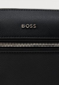 Μαύρη τσάντα από υφή δέρματος με ασημένιο φερμουάρ, με ένα λογότυπο που ξεχωρίζει πάνω από το φερμουάρ. Λεπτομερής ραφή γύρω από τις άκρες.