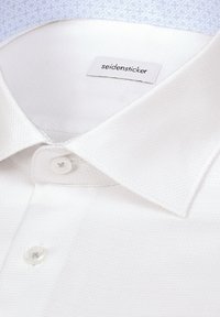 Gros plan d'un col de chemise blanche texturée avec deux boutons blancs et une étiquette de marque visible "seidensticker".