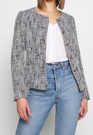 Vrouw draagt een zwart-wit gestructureerd jasje met knopen over een wit topje met stofbeklede knopen en lichtblauwe hooggetailleerde spijkerbroek.