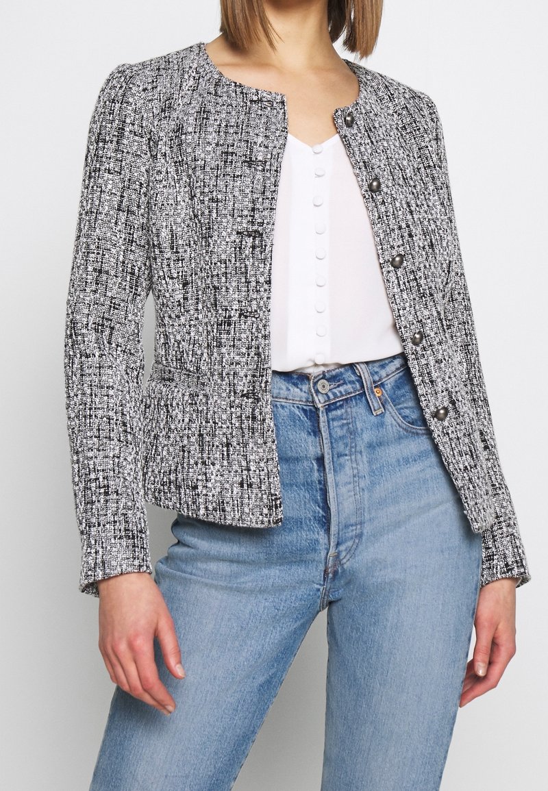Femme portant une veste boutonnée texturée noir et blanc sur un haut blanc avec des boutons recouverts de tissu et un jean taille haute bleu clair.