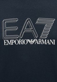Μπλε ναυτικό βαμβακερό μπλουζάκι με έντονο λευκό κείμενο: "EA7 EMPORIO ARMANI", με στυλιζαρισμένα γράμματα και διακριτικό λογότυπο αετού.