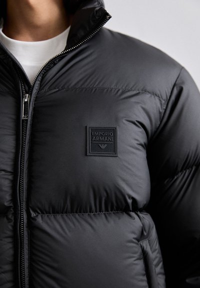 Doudoune noire avec un col haut, fermeture éclair et texture matelassée. Présente un patch logo noir texturé sur le côté gauche.