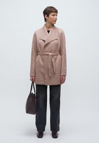 Ροζ ανοιχτό παλτό με ζώνη και γιακά σάλι, με δομημένο σχέδιο, συνδυασμένο με σκούρο τζιν και καφέ χειροποίητη τσάντα με υφή.