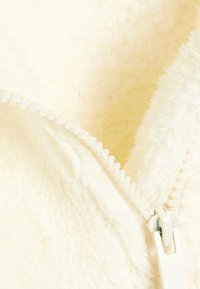 Tissu en polaire couleur crème avec une fermeture éclair visible. La texture est douce et moelleuse, avec un tirette de fermeture éclair lisse et de petites dents en plastique.
