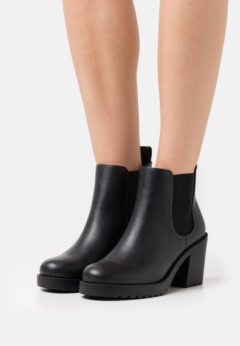 Bottes chevilles en cuir noir avec panneaux élastiques latéraux, talons carrés et languettes, portées sur des jambes nues sur un fond uni.