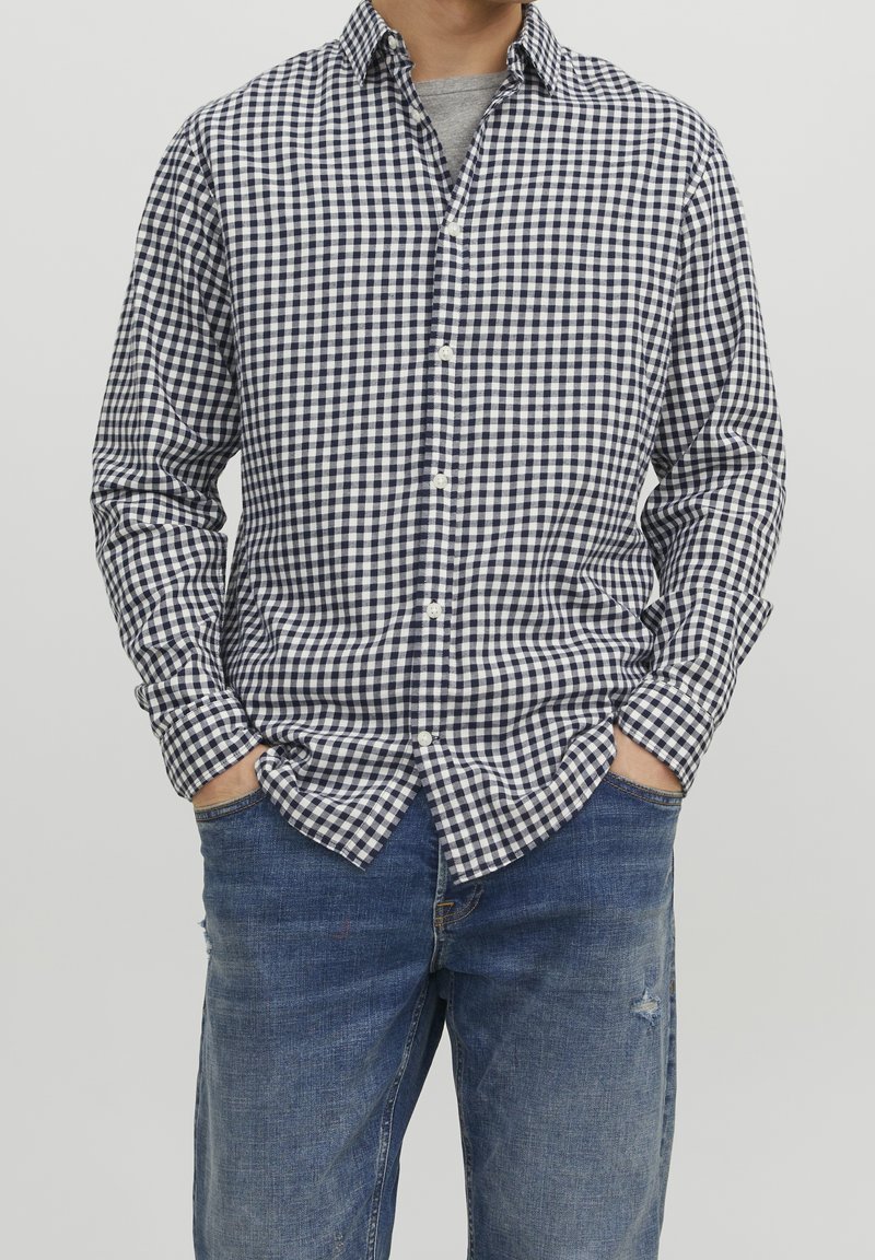 Camicia a quadri blu navy e bianca con bottoni, colletto, maniche lunghe e vestibilità comoda, abbinata a jeans blu con dettaglio usurato.