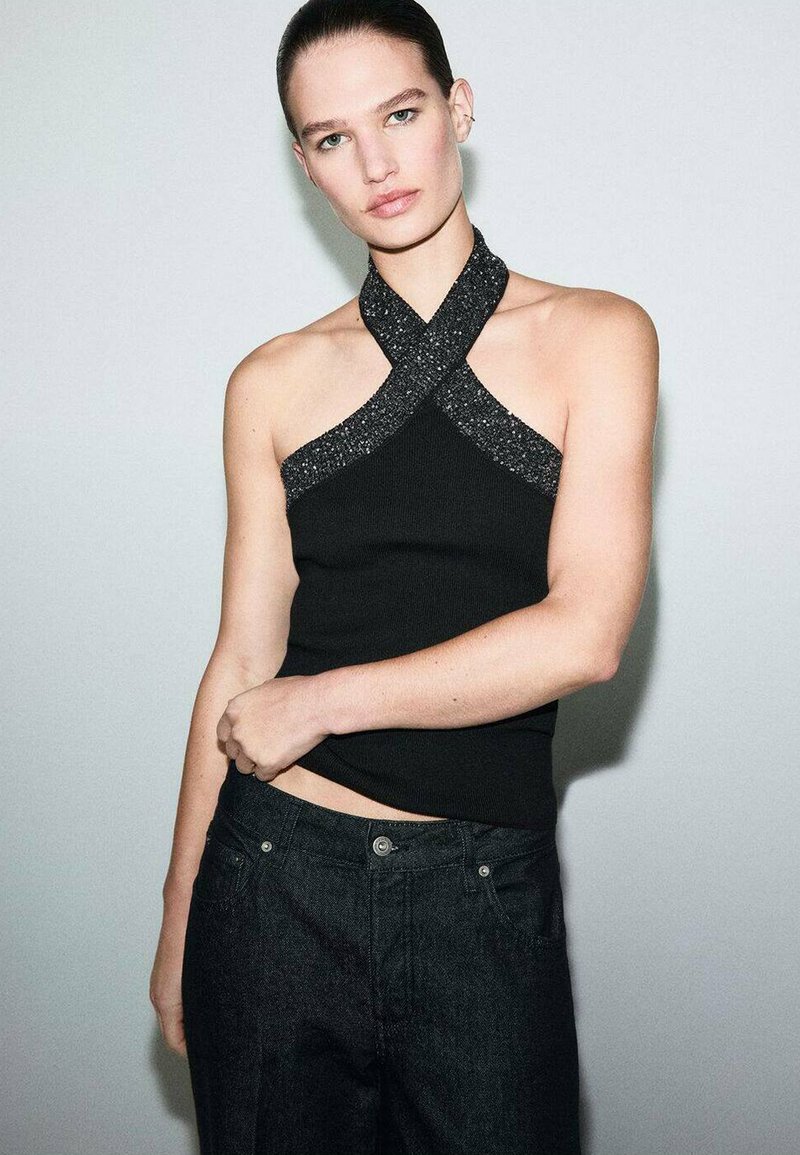 Top negro sin mangas con un diseño de tirantes cruzados que presenta adornos negros. Combinado con unos vaqueros oscursos, que exhiben una silueta ajustada.