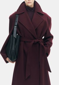 Bordeaux wollen jas met een brede revers, een ceintuur in de taille en oversized mouwen. Gecombineerd met een zwarte gestructureerde handtas.