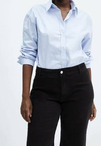 Persona con una camisa de botones de color azul claro, con las mangas enrolladas, metida dentro de pantalones negros de tiro alto, contra un fondo liso.