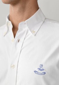 Primo piano di una persona che indossa una camicia bianca con bottoni e un logo Faconnable ricamato in blu sul petto.
