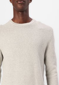 Gros plan sur le haut du torse d'une personne portant un pull en tricot beige clair avec un col rond sur un fond blanc.