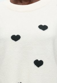 Gros plan d'un pull blanc avec des motifs de cœur tricotés noirs sur la poitrine et le torse.