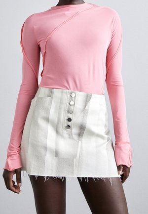 Dunkelhäutige Person, die ein pinkfarbenes Langarmshirt mit Daumenlöchern und einen weißes Minirock mit hoher Taille und silbernen Knöpfen trägt.